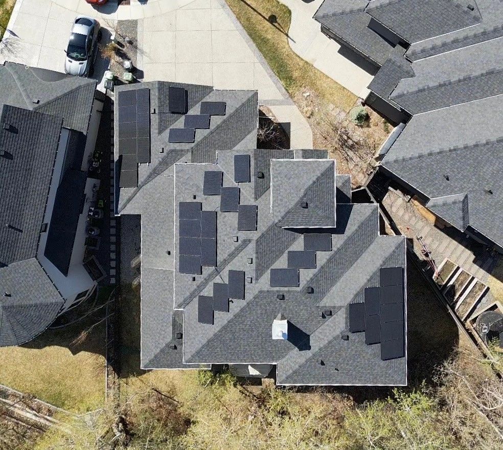 Aerial view of a complex residential roof with multiple levels and solar panels, surrounded by similar houses, a car, and a driveway.