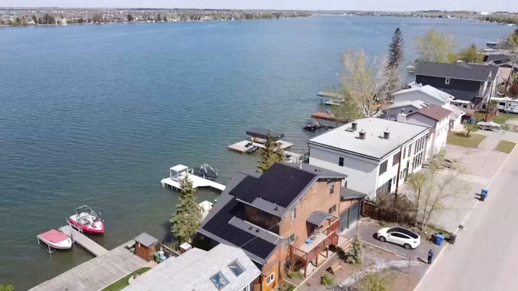 An aerial view of a lakeside residential area, with boats docked at private piers, houses nearby, and cars parked along a quiet street.