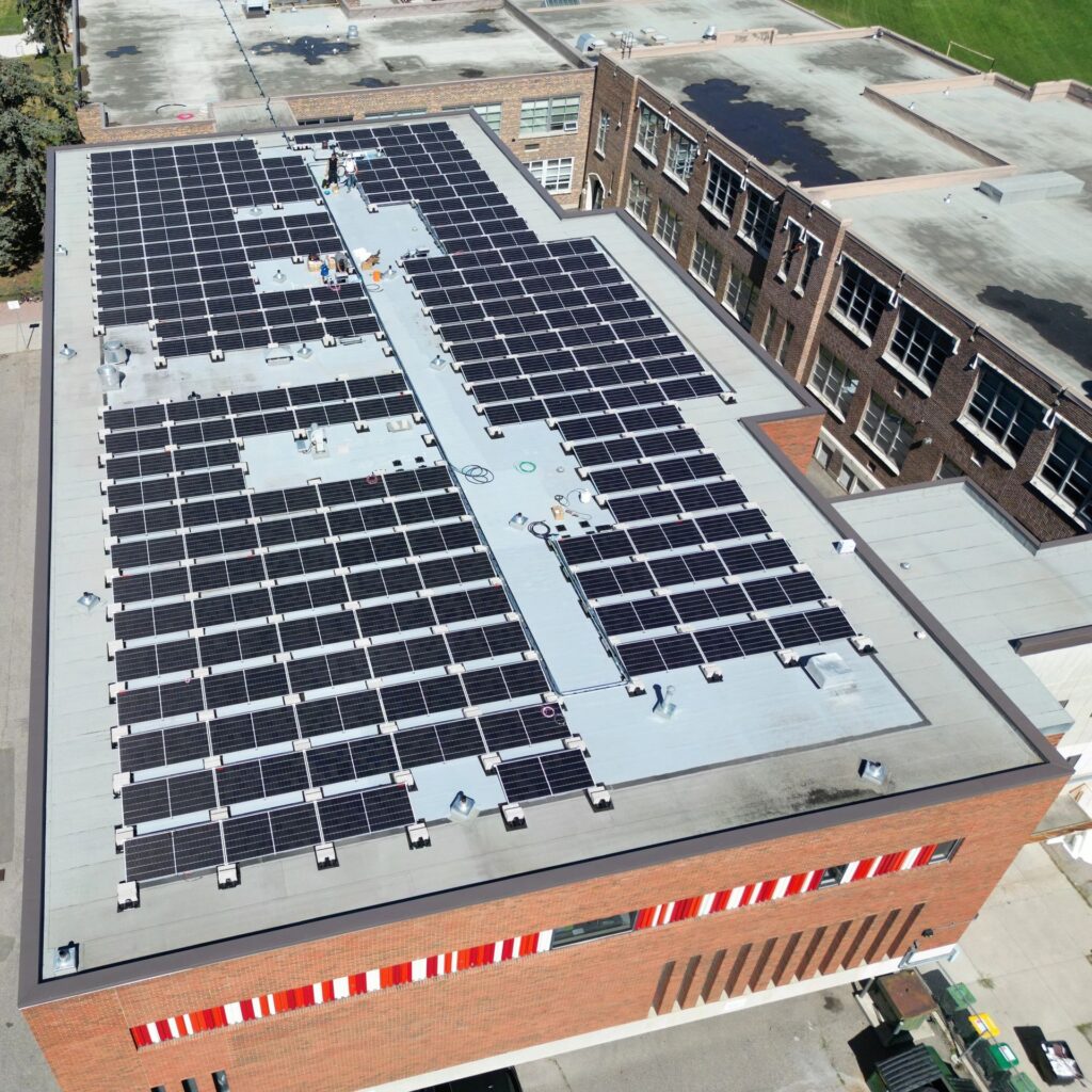 An aerial view of a brick building with a large solar panel array on the roof. The parking lot is partially empty, and it's a sunny day.