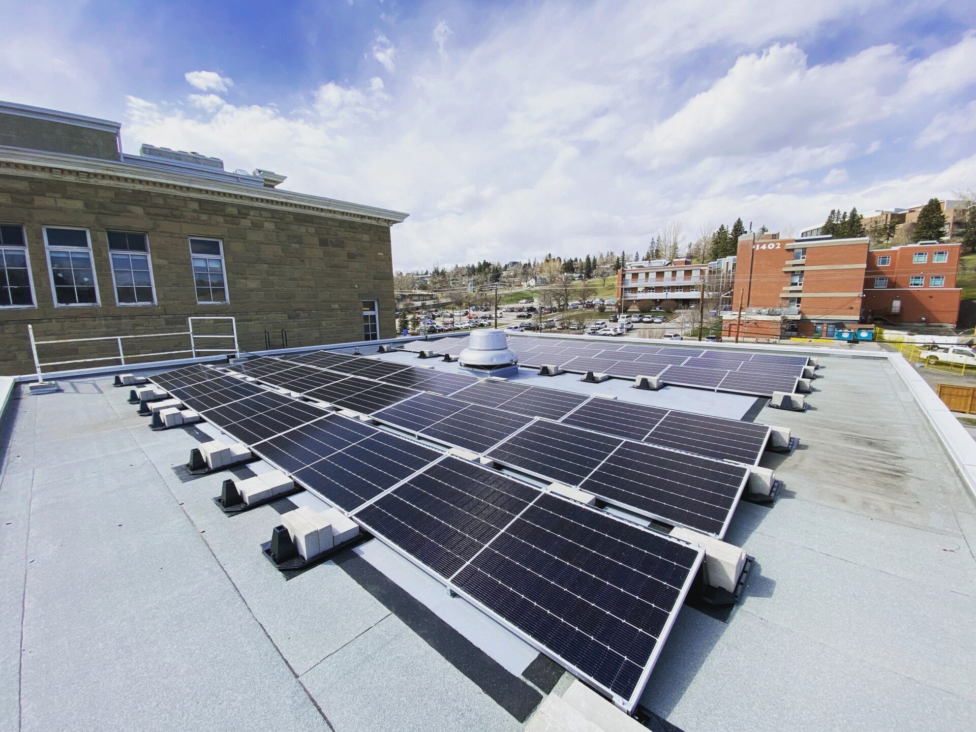 The image shows solar panels on a flat rooftop, with urban buildings in the background under a partly cloudy sky. It suggests renewable energy use in a city.