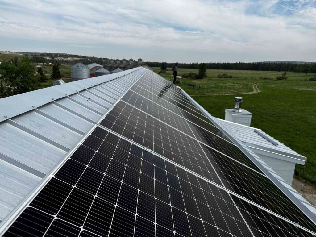 The image shows a series of solar panels installed on a slanted metal roof of a building, with agricultural silo structures and open countryside in the background.