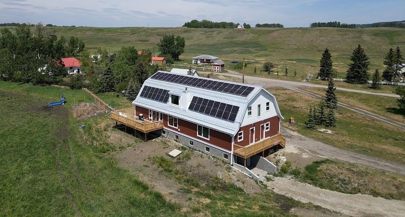 Aerial view of a two-story house with a solar panel array on the roof. Surrounded by greenery, a deck, and rural landscape with scattered buildings.