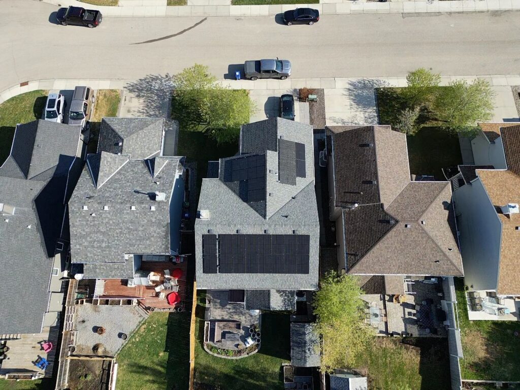 An aerial view of a suburban neighborhood showing rooftops, some with solar panels, cars parked on a street, and backyards with trees and lawns.