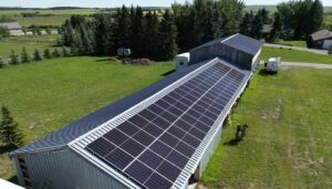 An aerial view of a large building with a roof covered in solar panels surrounded by green grass, trees, and a clear blue sky above.