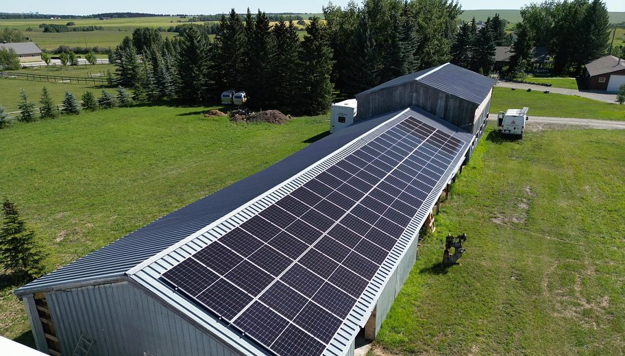 An aerial view of a large building with a roof covered in solar panels surrounded by green grass, trees, and a clear blue sky above.
