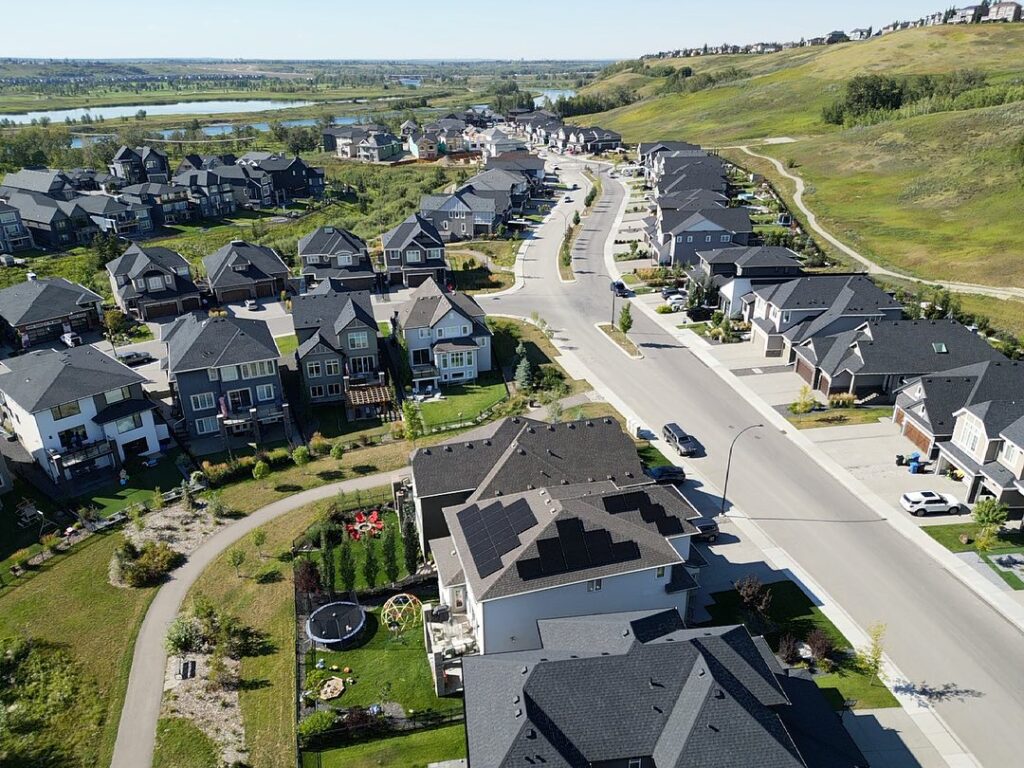 Aerial view of a suburban neighborhood with neat rows of houses, manicured lawns, a winding road, and a distant lake with greenery.
