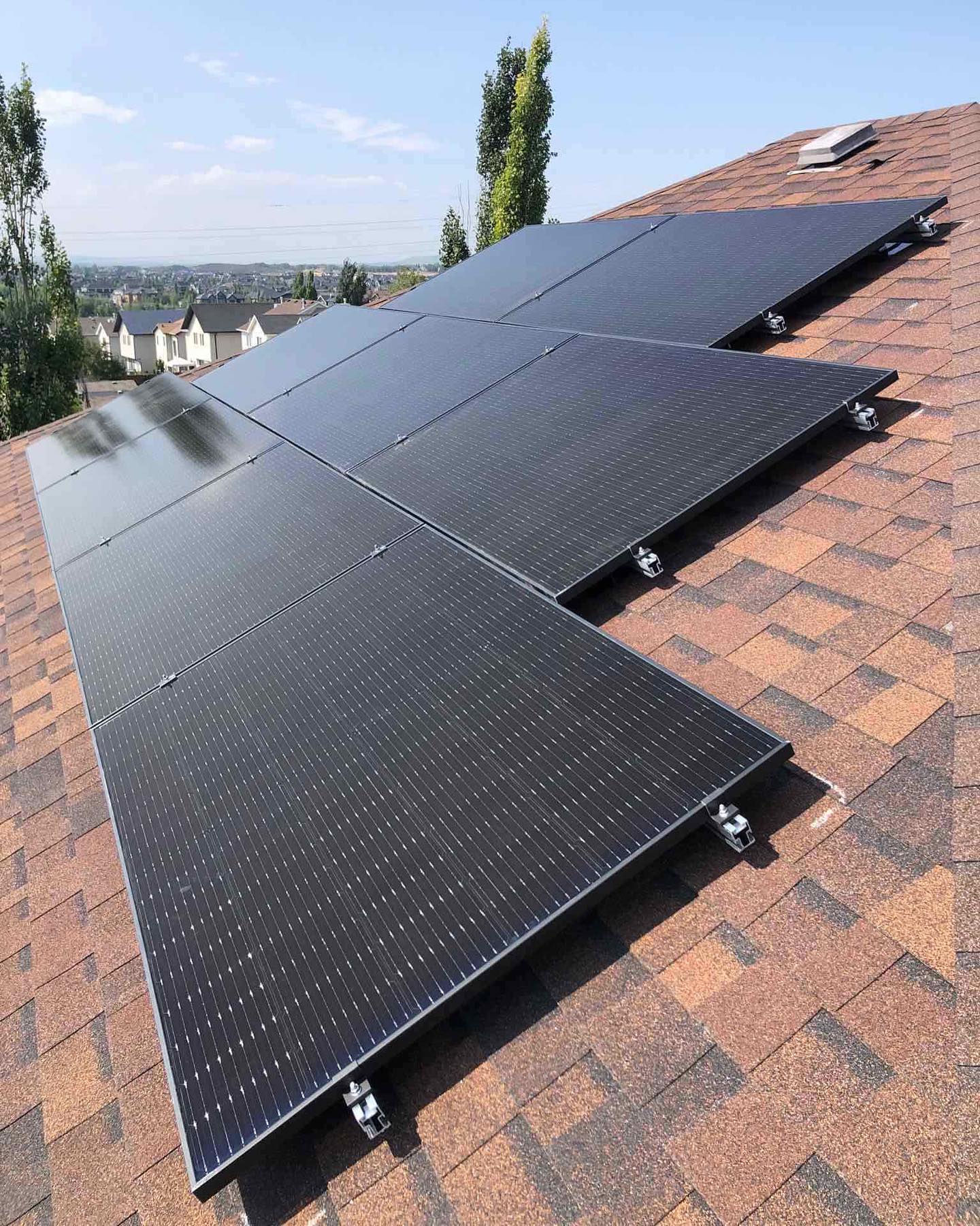 This image shows a series of solar panels installed on the shingled roof of a house with a suburban landscape and clear sky in the background.