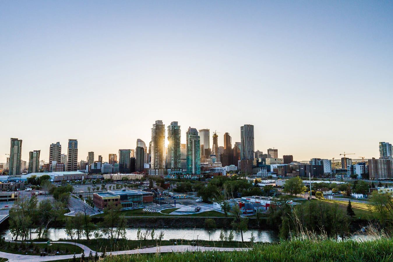 A city skyline at sunset with the sun reflecting off high-rise buildings, a river in the foreground, and a clear sky above.