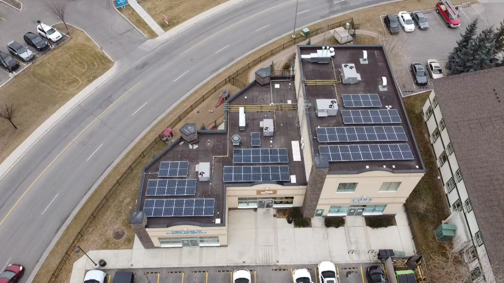 This is an aerial view of a two-story building complex with solar panels on the roofs, adjacent to a road, with parked cars alongside.