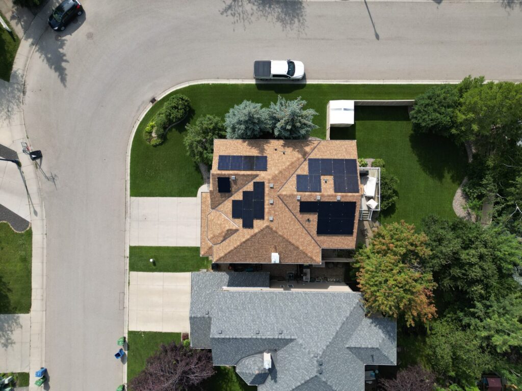 Aerial view of a residential area with a house featuring solar panels on its roof, surrounded by green lawns, trees, and a cul-de-sac street.
