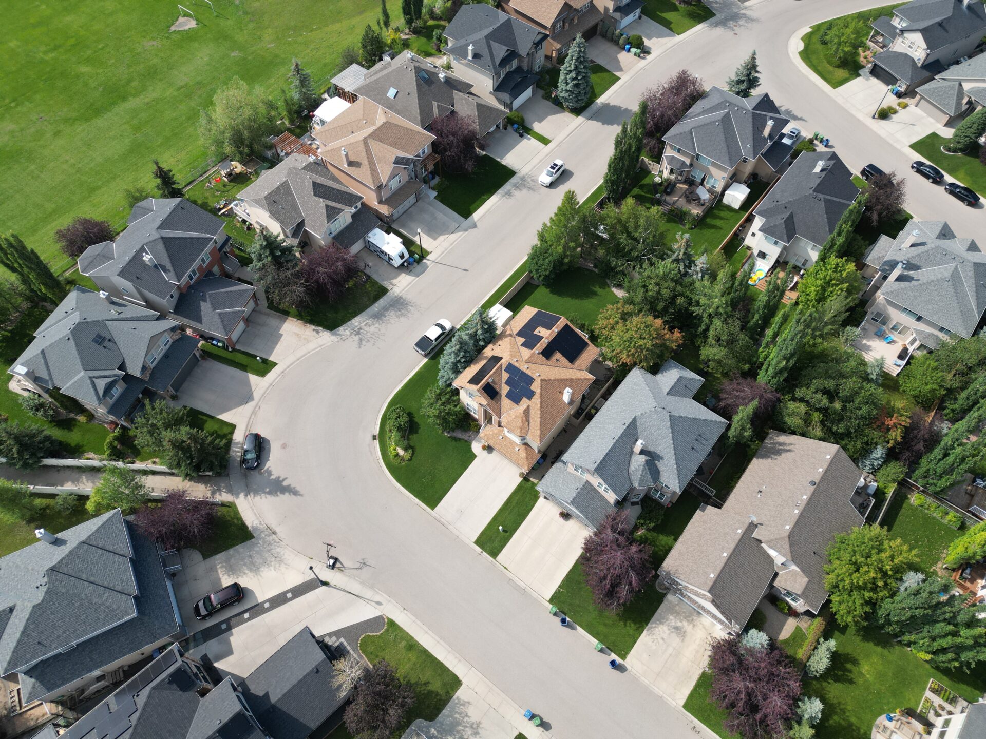 Aerial view of a residential neighborhood with several detached houses, manicured lawns, trees, vehicles, and curving streets on a sunny day.