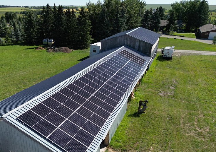 This aerial image shows a large barn with a solar panel-covered roof amidst green surroundings, with vehicles parked nearby and trees in the background.
