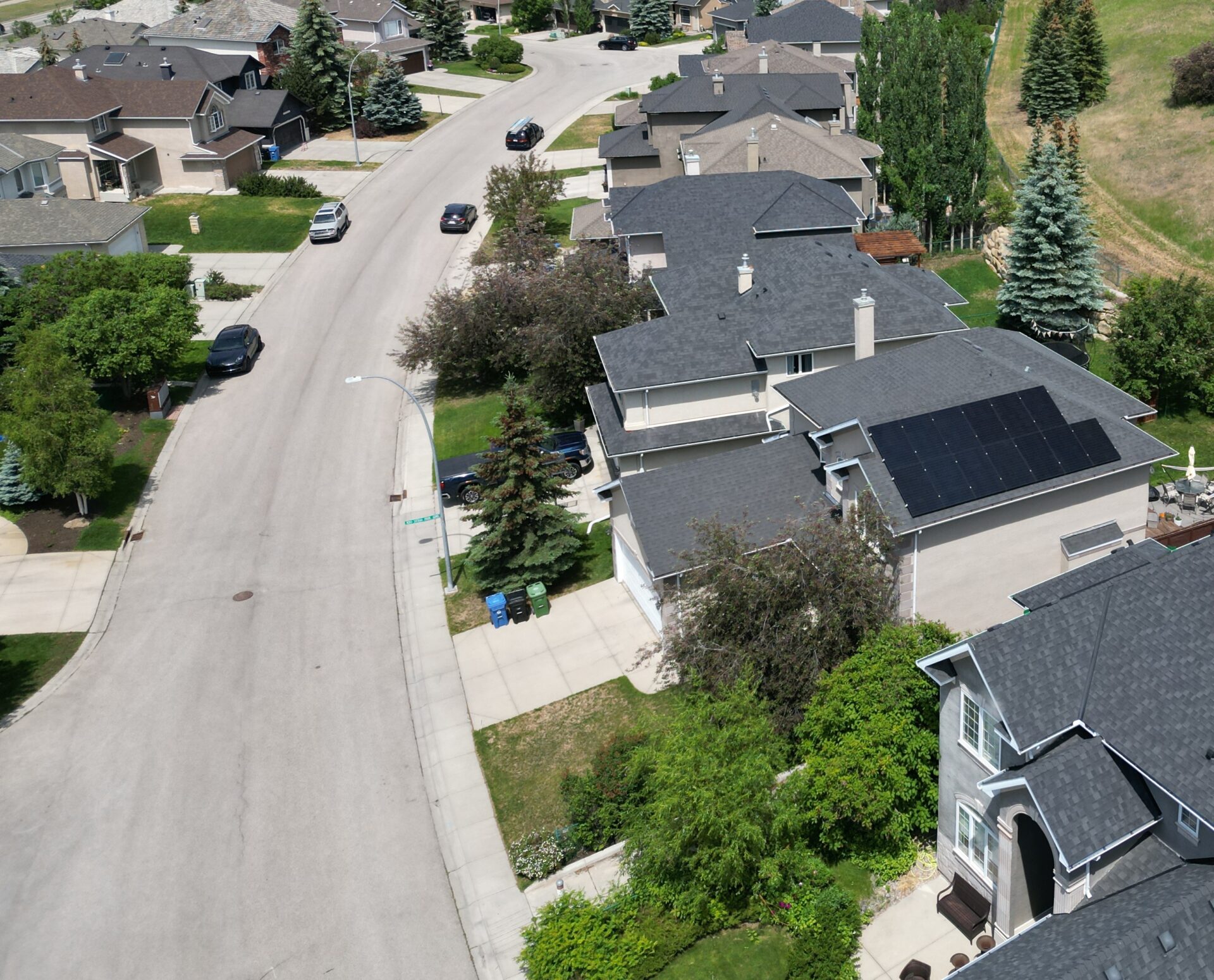 An aerial view of a suburban neighborhood with houses, solar panels on a roof, lush trees, green lawns, and cars on the street.