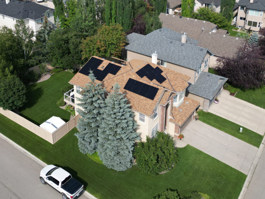 Aerial view of a suburban two-story house with a manicured lawn, solar panels on the roof, surrounded by trees, with a SUV parked outside.
