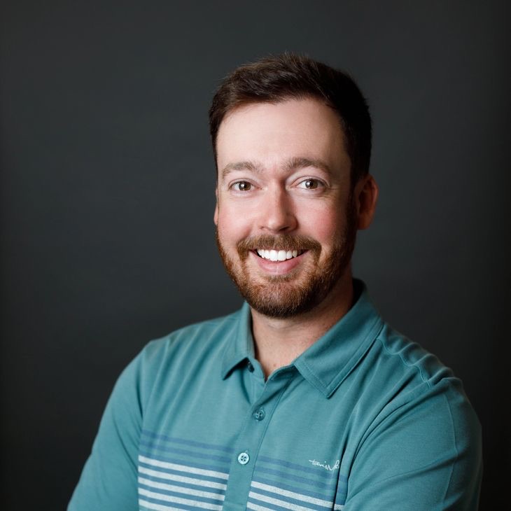 A smiling person with short hair and a beard, wearing a striped polo shirt, is photographed against a plain dark background.