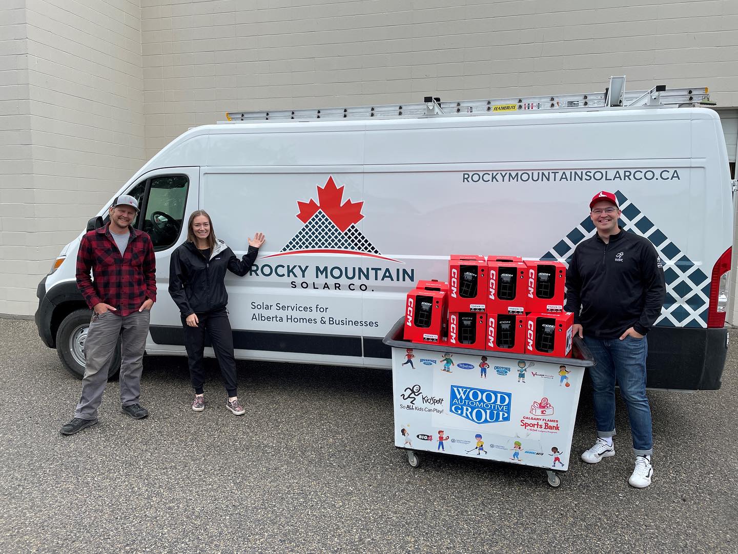 Three people stand smiling beside a white van with "ROCKY MOUNTAIN SOLAR CO." branding, and a cart loaded with red portable generators.