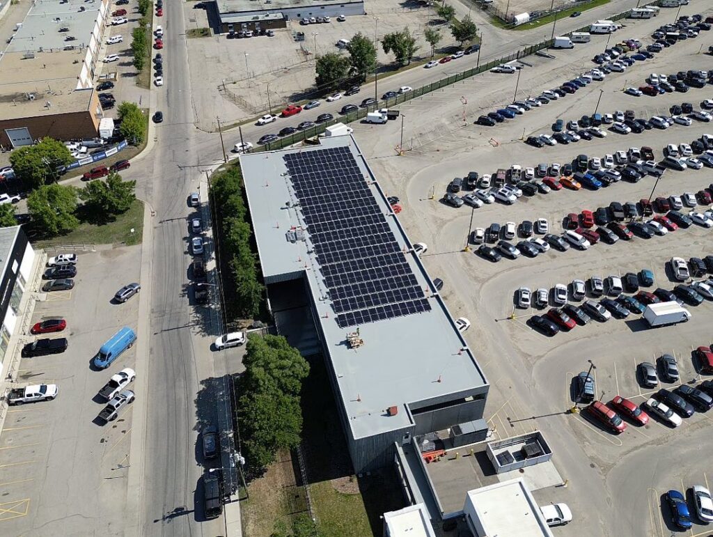 Aerial view of a building with solar panels on the roof, surrounded by parked cars in a large lot, on a sunny day with clear skies.
