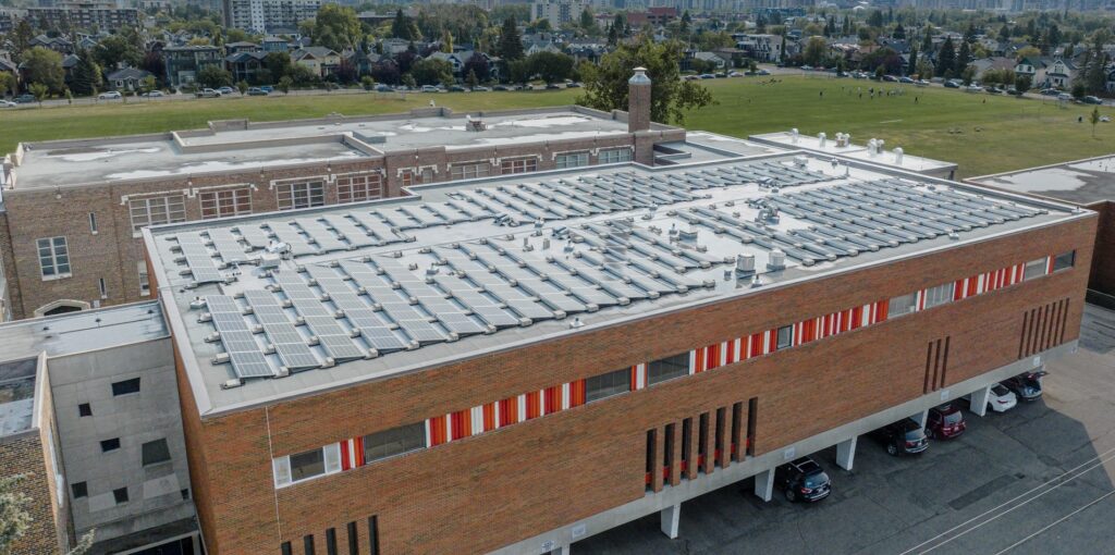 Aerial view of a large brick building with a flat roof covered in rows of solar panels. Cars are parked under an overhang, adjacent to a large field.