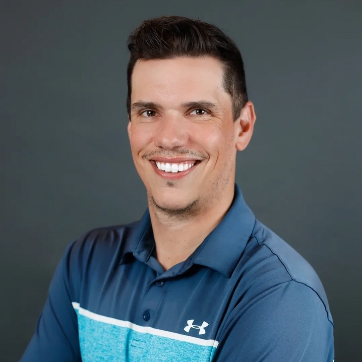 A person with short dark hair, a friendly smile, wearing a blue Under Armour shirt, posing in front of a grey background.