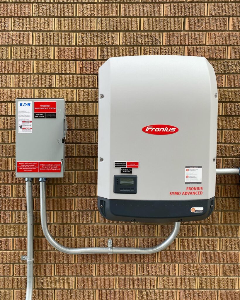 A Fronius inverter and electrical disconnect box mounted on a brick wall, connected with metal conduit, part of a photovoltaic solar installation.