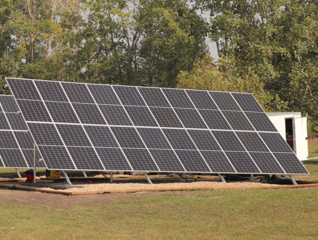 A large solar panel array is installed in a grassy field with trees in the background and a small structure nearby.