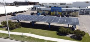A large solar panel array forms a canopy above parking spaces in front of a Chevrolet Buick GMC dealership, with clear skies above.