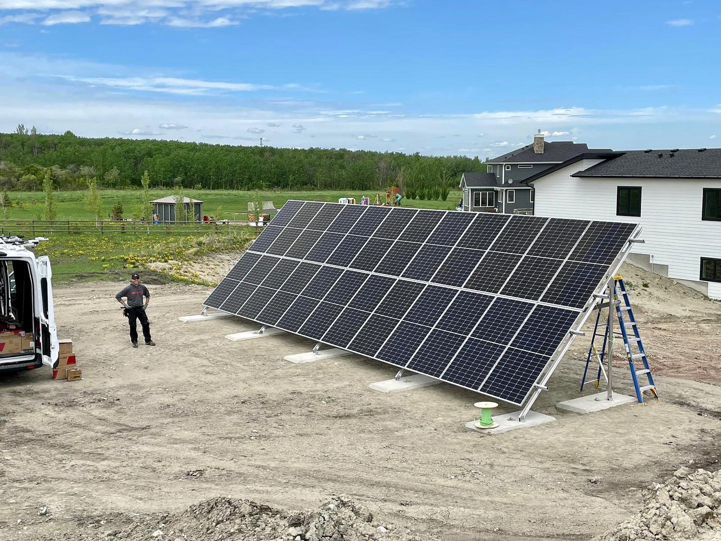 A person stands by a large solar panel array next to a house under a clear sky, with a ladder and open van showing a work scene.
