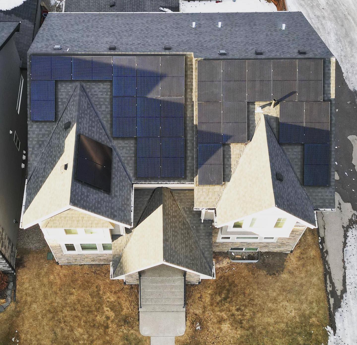 Aerial view of a large house with multiple roof sections, several equipped with solar panels. Snow patches are visible around the property, indicating winter.