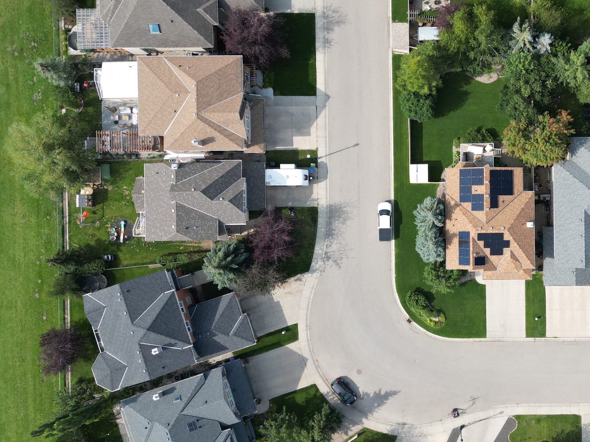 Aerial view of a residential neighborhood with houses, lush green lawns, driveways, vehicles, and a curved street. Solar panels visible on one roof.