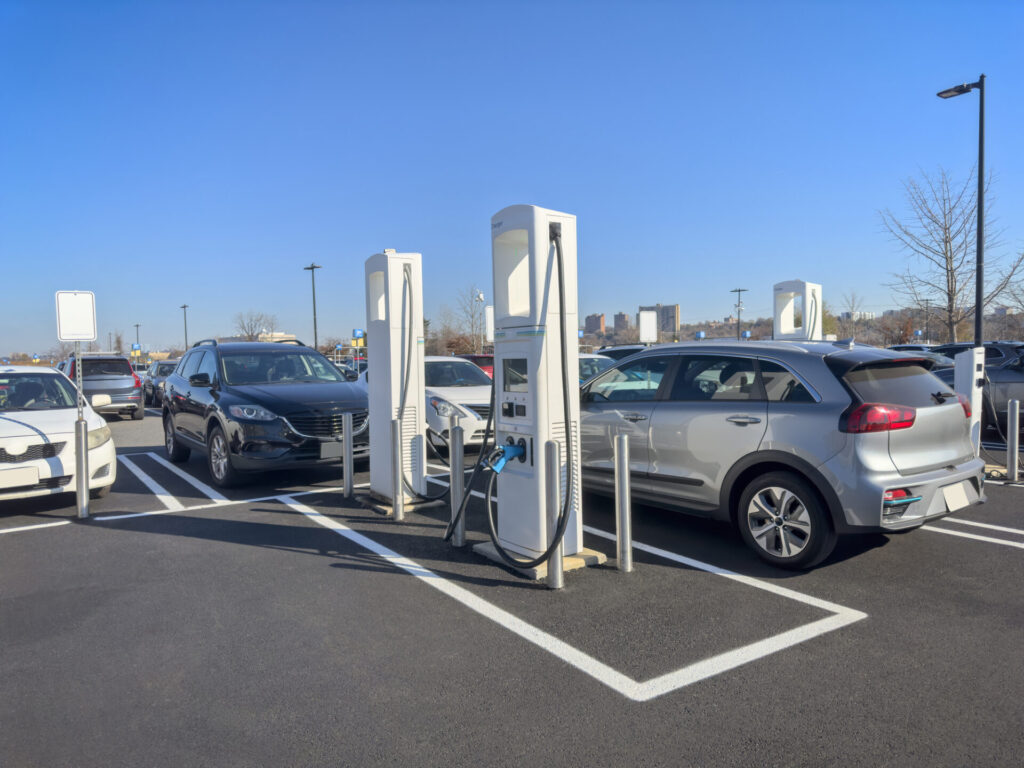 A parking lot with electric vehicle charging stations and cars plugged in. Clear blue sky above, with urban buildings in the background.
