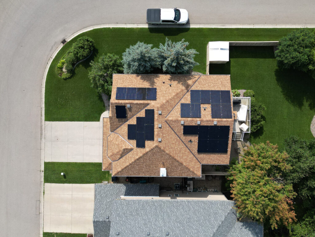 Aerial view of a suburban house with solar panels on the roof, a neatly manicured lawn, a white car parked out front, and adjacent houses.