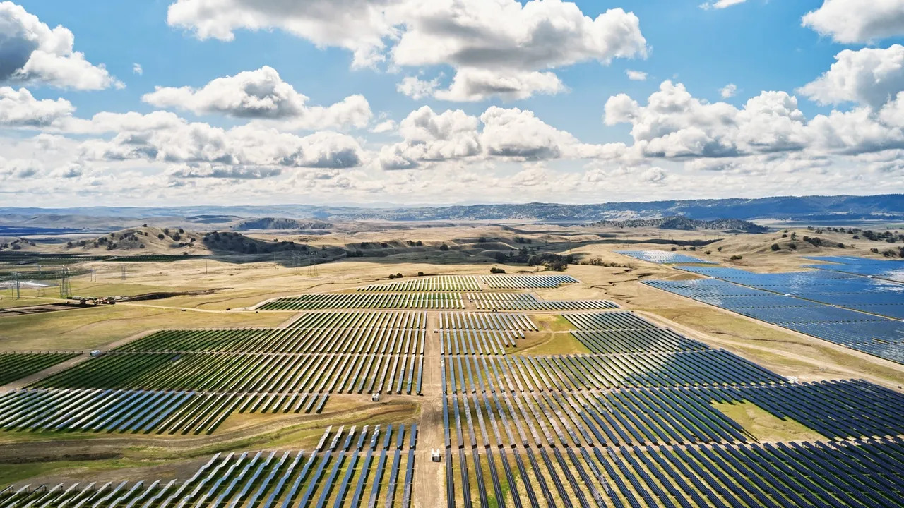 A vast solar farm stretches across rolling hills under a dynamic sky with clouds casting shadows over the panels and the surrounding countryside.