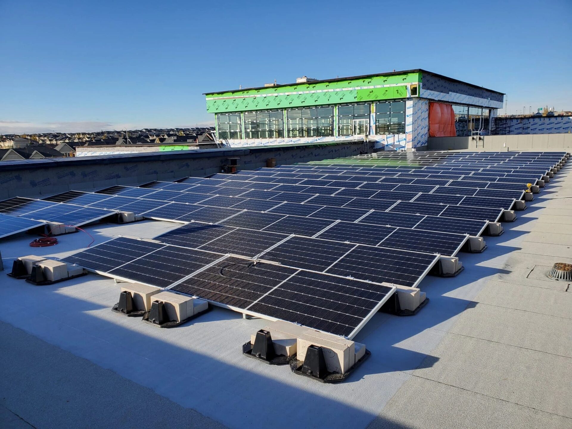 Rows of solar panels installed on a flat rooftop with a building under construction in the background under a clear blue sky.