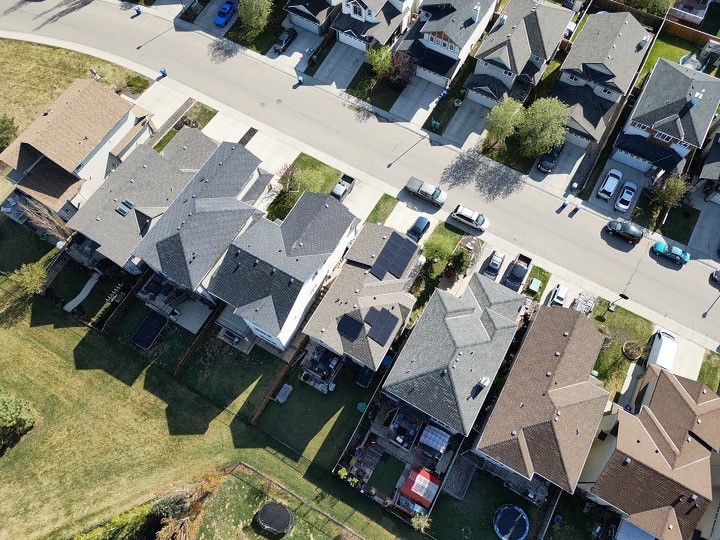 An aerial view of a suburban neighborhood with houses, backyards, vehicles parked on streets and driveways, and green lawns on a sunny day.