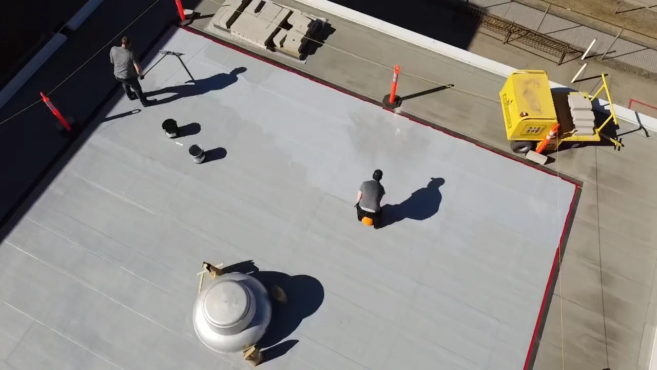 Aerial view of two people working on a rooftop near HVAC units and a maintenance cart, with visible shadows and safety cones demarcating the area.