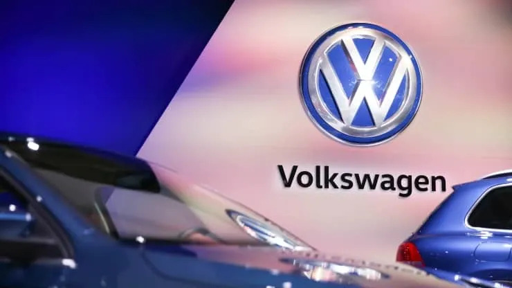 This image shows the Volkswagen logo illuminated above a model name on a smooth backdrop, with the partial reflection of a blue car visible.