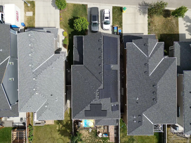 An aerial view of a residential area with houses featuring dark roofs, neat yards, and parked vehicles along a tree-lined street.