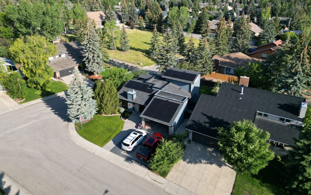 Aerial view of a residential neighborhood with detached houses, mature trees, green lawns, solar panels on roofs, cars parked on driveways, and a cul-de-sac.