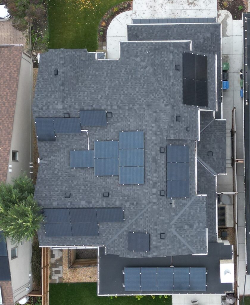 Aerial view of a gray-roofed building with multiple solar panels, surrounded by a green lawn, driveway, and a neighboring house.