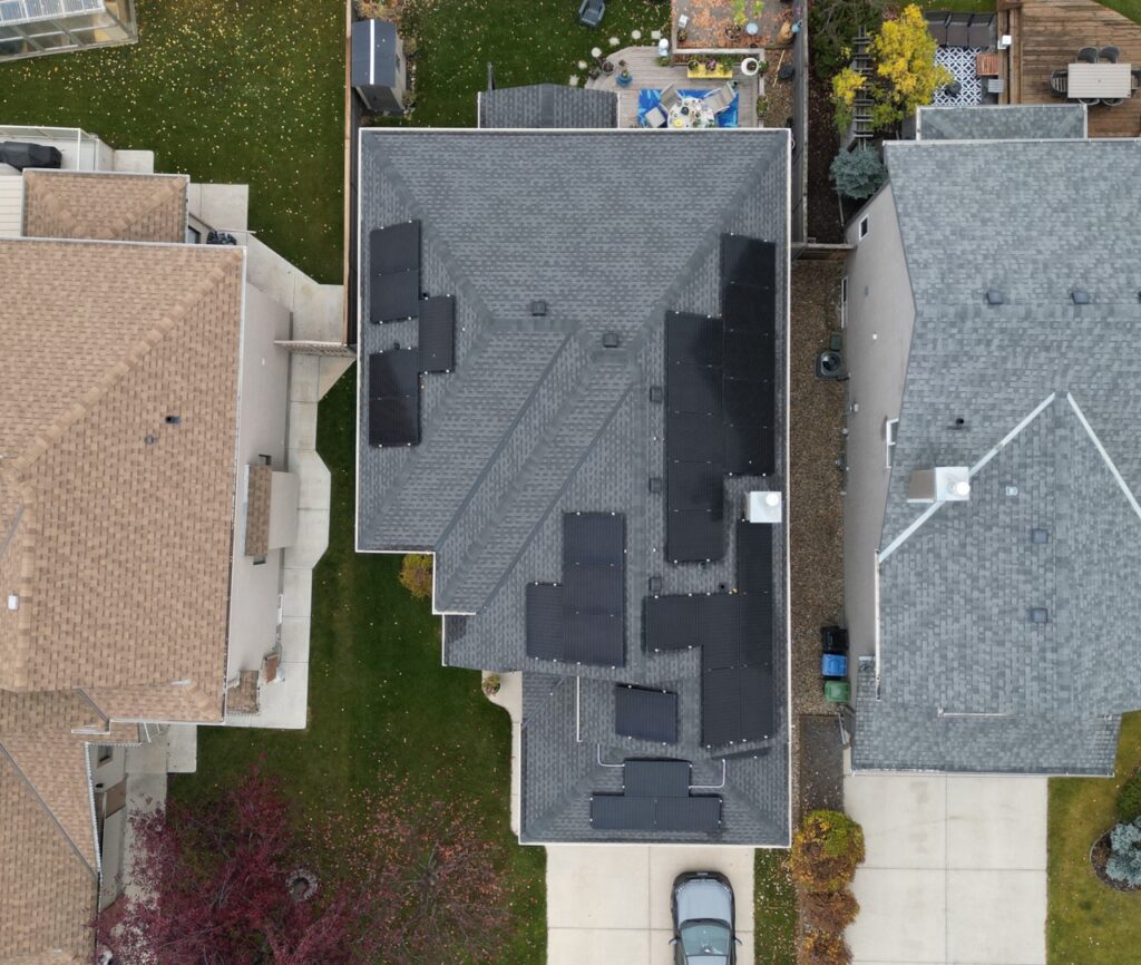 Aerial view of a residential area showing the tops of houses with varying roof designs, surrounded by lawns, trees, and a driveway with vehicles.