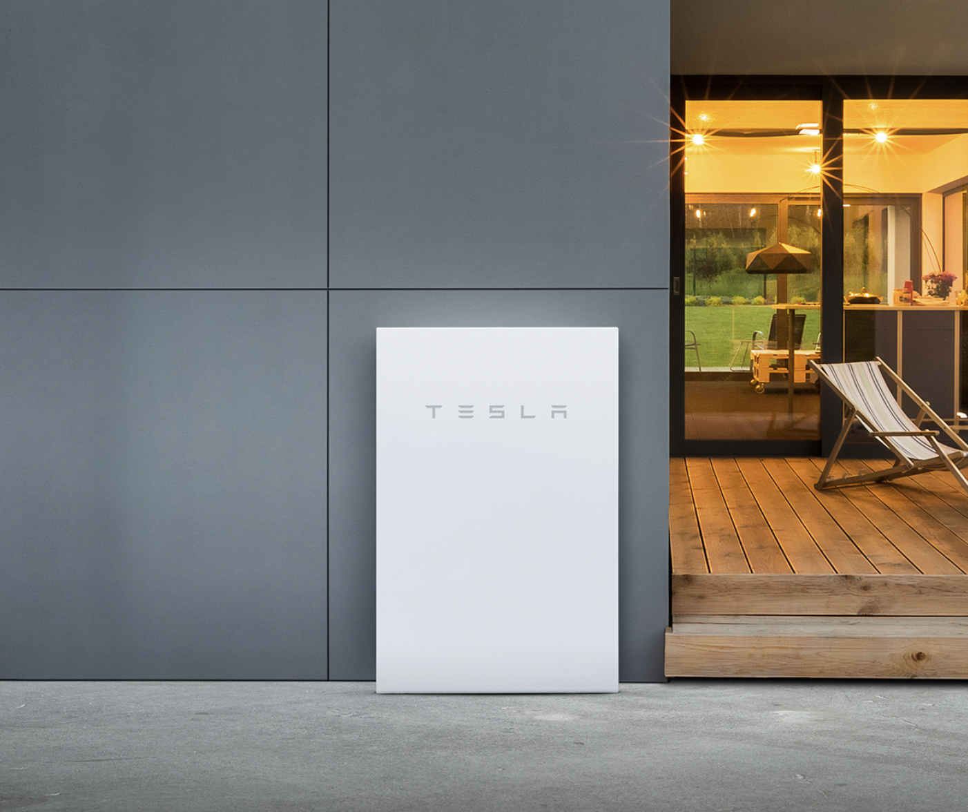 A white Tesla Powerwall battery is mounted on a gray wall outside a modern house with visible interior lights and a wooden deck with a chair.